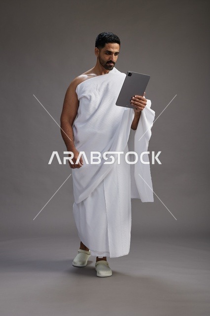 A portrait of a Saudi pilgrim in Ihram clothing holding a tablet, preparing to perform the rituals of Hajj and Umrah, full body, gray background.