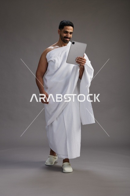 A portrait of a Saudi pilgrim in Ihram clothing holding a tablet, preparing to perform the rituals of Hajj and Umrah, full body, gray background.