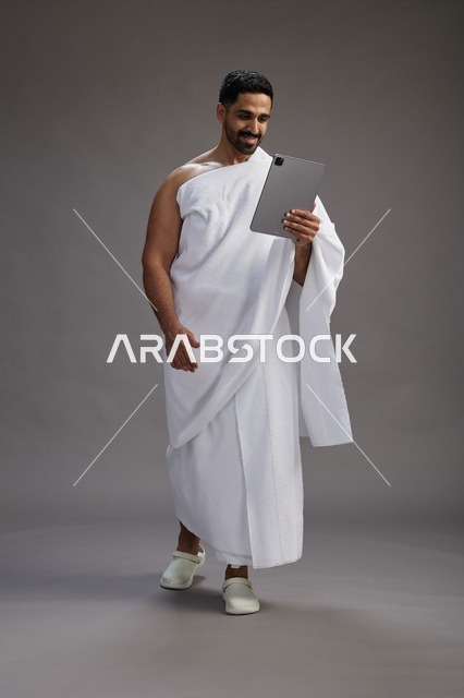 بورتريه لحاج سعودي بملابس الإحرام يحمل تابلت, الاستعداد لاداء مناسك الحج والعمره, كامل الجسم, خلفيه رماديه