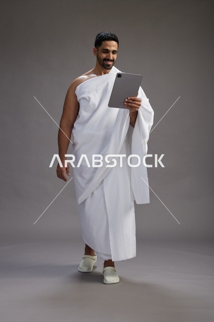بورتريه لحاج سعودي بملابس الإحرام يحمل تابلت, الاستعداد لاداء مناسك الحج والعمره, كامل الجسم, خلفيه رماديه