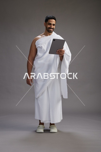 A portrait of a Saudi pilgrim in Ihram clothing holding a tablet, preparing to perform the rituals of Hajj and Umrah, full body, gray background.