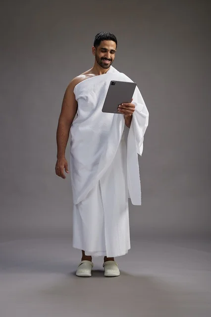 A portrait of a Saudi pilgrim in Ihram clothing holding a tablet, preparing to perform the rituals of Hajj and Umrah, full body, gray background.