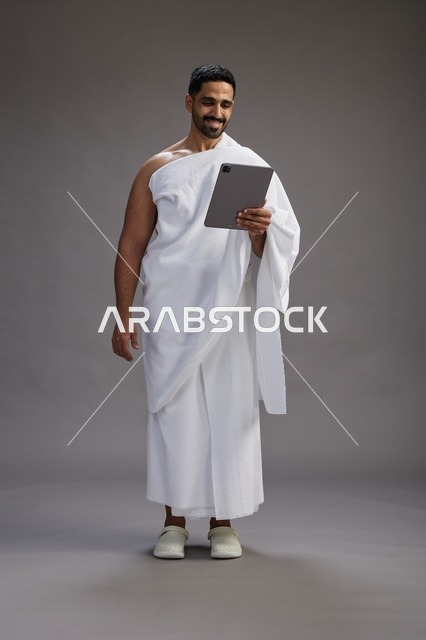 بورتريه لحاج سعودي بملابس الإحرام يحمل تابلت, الاستعداد لاداء مناسك الحج والعمره, كامل الجسم, خلفيه رماديه
