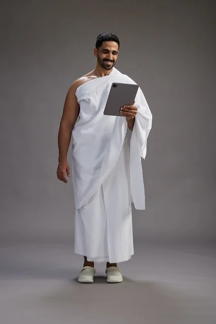A portrait of a Saudi pilgrim in Ihram clothing holding a tablet, preparing to perform the rituals of Hajj and Umrah, full body, gray background.