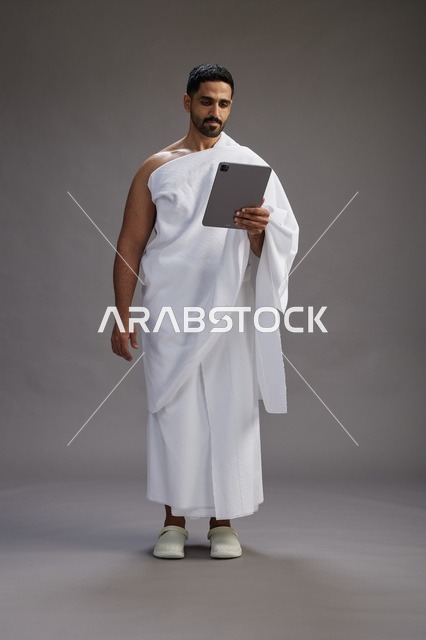 بورتريه لحاج سعودي بملابس الإحرام يحمل تابلت, الاستعداد لاداء مناسك الحج والعمره, كامل الجسم, خلفيه رماديه