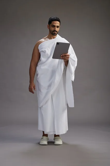 A portrait of a Saudi pilgrim in Ihram clothing holding a tablet, preparing to perform the rituals of Hajj and Umrah, full body, gray background.