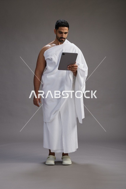 بورتريه لحاج سعودي بملابس الإحرام يحمل تابلت, الاستعداد لاداء مناسك الحج والعمره, كامل الجسم, خلفيه رماديه