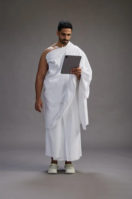 A portrait of a Saudi pilgrim in Ihram clothing holding a tablet, preparing to perform the rituals of Hajj and Umrah, full body, gray background.