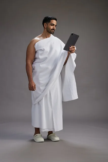 A portrait of a Saudi pilgrim in Ihram clothing holding a tablet, preparing to perform the rituals of Hajj and Umrah, full body, gray background.