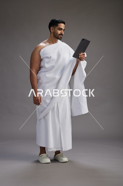 A portrait of a Saudi pilgrim in Ihram clothing holding a tablet, preparing to perform the rituals of Hajj and Umrah, full body, gray background.