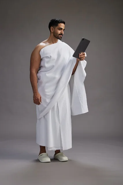 A portrait of a Saudi pilgrim in Ihram clothing holding a tablet, preparing to perform the rituals of Hajj and Umrah, full body, gray background.