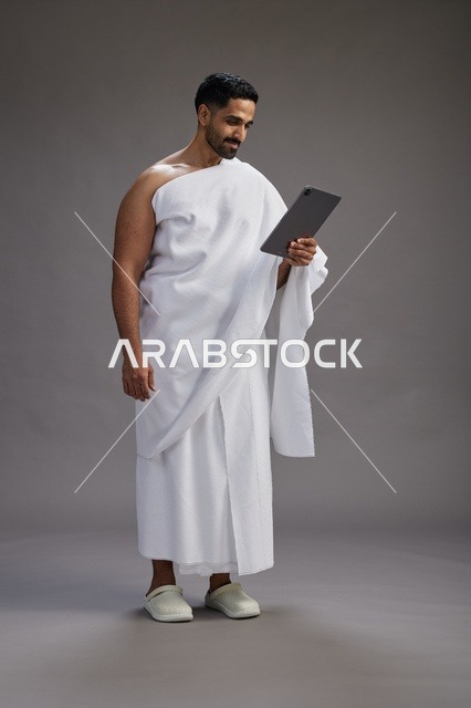 بورتريه لحاج سعودي بملابس الإحرام يحمل تابلت, الاستعداد لاداء مناسك الحج والعمره, كامل الجسم, خلفيه رماديه