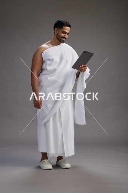 بورتريه لحاج سعودي بملابس الإحرام يحمل تابلت, الاستعداد لاداء مناسك الحج والعمره, كامل الجسم, خلفيه رماديه