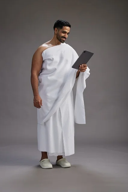 بورتريه لحاج سعودي بملابس الإحرام يحمل تابلت, الاستعداد لاداء مناسك الحج والعمره, كامل الجسم, خلفيه رماديه