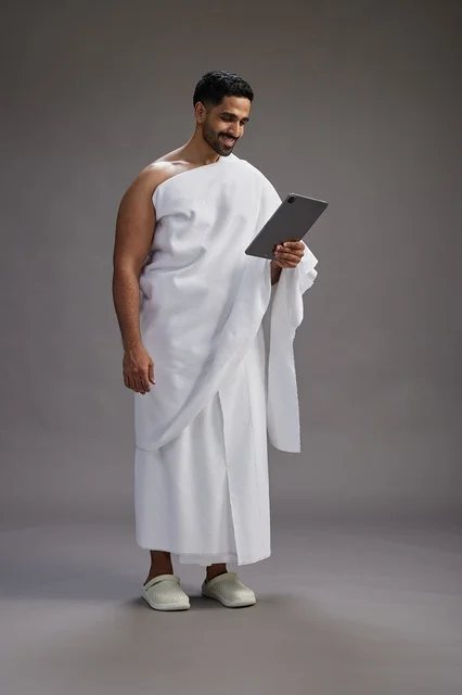 A portrait of a Saudi pilgrim in Ihram clothing holding a tablet, preparing to perform the rituals of Hajj and Umrah, full body, gray background.