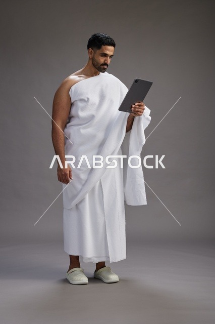 بورتريه لحاج سعودي بملابس الإحرام يحمل تابلت, الاستعداد لاداء مناسك الحج والعمره, كامل الجسم, خلفيه رماديه
