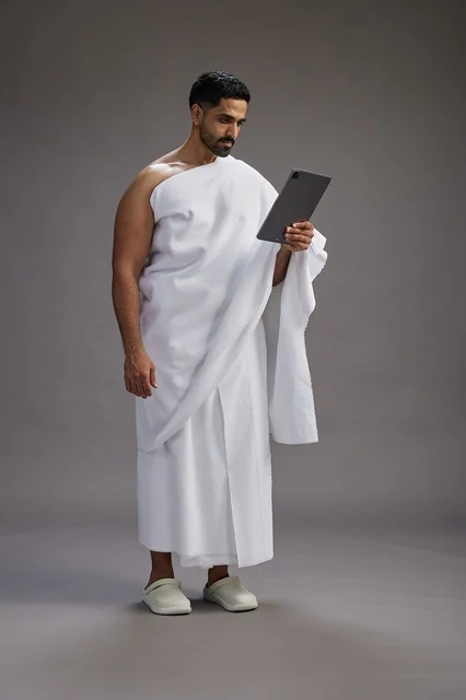A portrait of a Saudi pilgrim in Ihram clothing holding a tablet, preparing to perform the rituals of Hajj and Umrah, full body, gray background.