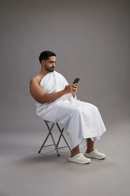 A portrait of a Saudi pilgrim in Ihram clothing using a mobile phone and sitting on a chair, preparing to perform the rituals of Hajj and Umrah, with a gray background.