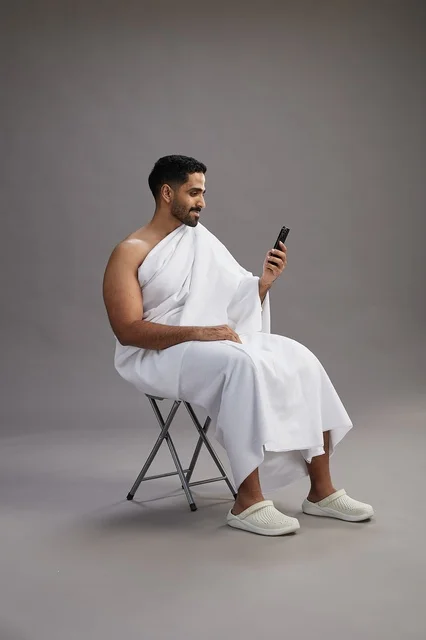 A portrait of a Saudi pilgrim in Ihram clothing using a mobile phone and sitting on a chair, preparing to perform the rituals of Hajj and Umrah, with a gray background.