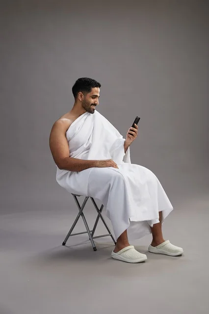 A portrait of a Saudi pilgrim in Ihram clothing using a mobile phone and sitting on a chair, preparing to perform the rituals of Hajj and Umrah, with a gray background.