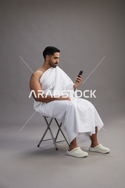 بورتريه لحاج سعودي بملابس الإحرام يستخدم الهاتف المحمول ويجلس على كرسي, الاستعداد لاداء مناسك الحج والعمره, خلفيه رماديه