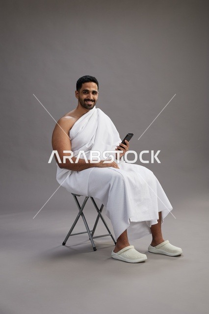 A portrait of a Saudi pilgrim in Ihram clothing using a mobile phone and sitting on a chair, preparing to perform the rituals of Hajj and Umrah, with a gray background.