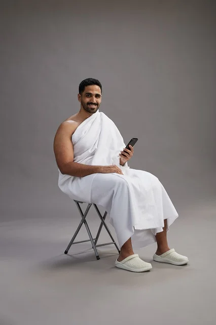 A portrait of a Saudi pilgrim in Ihram clothing using a mobile phone and sitting on a chair, preparing to perform the rituals of Hajj and Umrah, with a gray background.