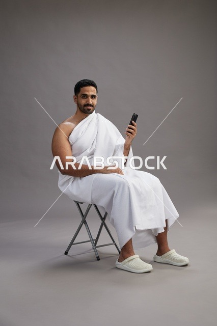 A portrait of a Saudi pilgrim in Ihram clothing using a mobile phone and sitting on a chair, preparing to perform the rituals of Hajj and Umrah, with a gray background.