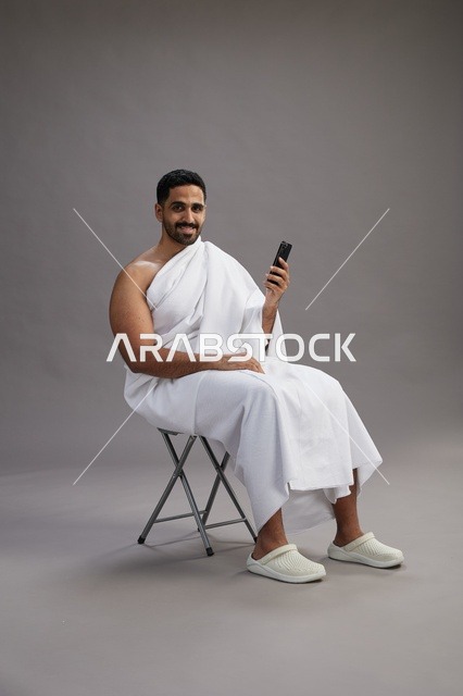 بورتريه لحاج سعودي بملابس الإحرام يستخدم الهاتف المحمول ويجلس على كرسي, الاستعداد لاداء مناسك الحج والعمره, خلفيه رماديه