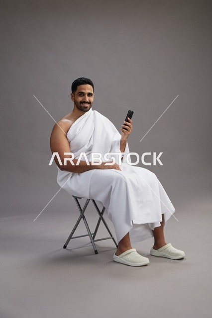 بورتريه لحاج سعودي بملابس الإحرام يستخدم الهاتف المحمول ويجلس على كرسي, الاستعداد لاداء مناسك الحج والعمره, خلفيه رماديه
