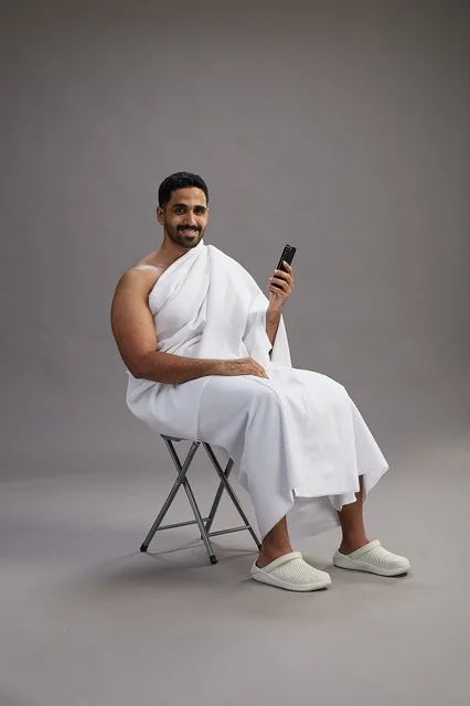 بورتريه لحاج سعودي بملابس الإحرام يستخدم الهاتف المحمول ويجلس على كرسي, الاستعداد لاداء مناسك الحج والعمره, خلفيه رماديه