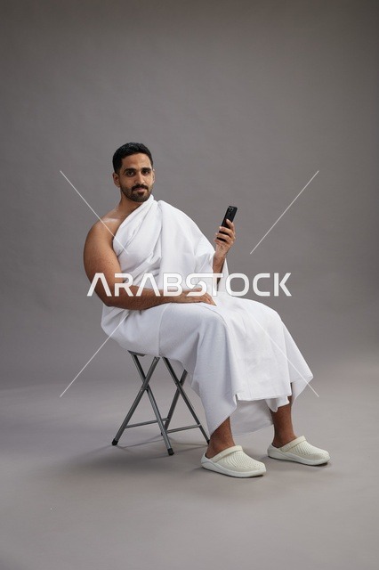 بورتريه لحاج سعودي بملابس الإحرام يستخدم الهاتف المحمول ويجلس على كرسي, الاستعداد لاداء مناسك الحج والعمره, خلفيه رماديه