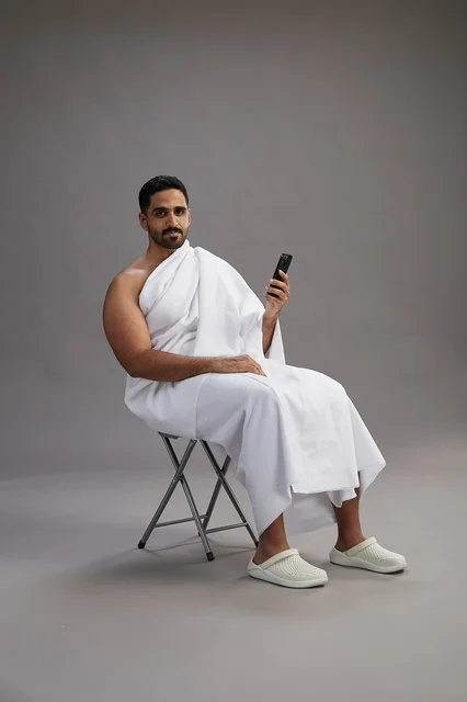 بورتريه لحاج سعودي بملابس الإحرام يستخدم الهاتف المحمول ويجلس على كرسي, الاستعداد لاداء مناسك الحج والعمره, خلفيه رماديه