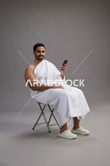 بورتريه لحاج سعودي بملابس الإحرام يستخدم الهاتف المحمول ويجلس على كرسي, الاستعداد لاداء مناسك الحج والعمره, خلفيه رماديه