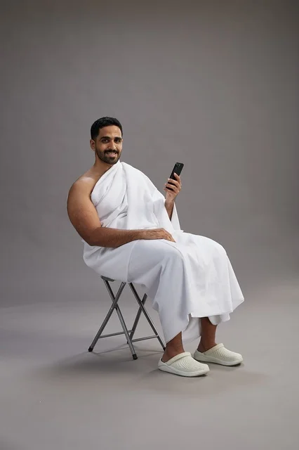 A portrait of a Saudi pilgrim in Ihram clothing using a mobile phone and sitting on a chair, preparing to perform the rituals of Hajj and Umrah, with a gray background.