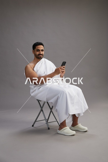 A portrait of a Saudi pilgrim in Ihram clothing using a mobile phone and sitting on a chair, preparing to perform the rituals of Hajj and Umrah, with a gray background.