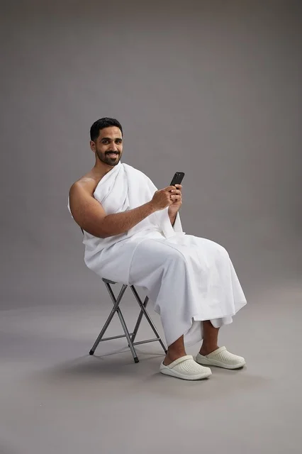 A portrait of a Saudi pilgrim in Ihram clothing using a mobile phone and sitting on a chair, preparing to perform the rituals of Hajj and Umrah, with a gray background.