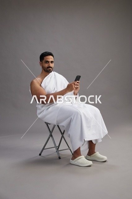 A portrait of a Saudi pilgrim in Ihram clothing using a mobile phone and sitting on a chair, preparing to perform the rituals of Hajj and Umrah, with a gray background.