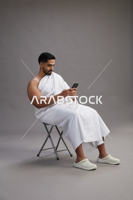 بورتريه لحاج سعودي بملابس الإحرام يستخدم الهاتف المحمول ويجلس على كرسي, الاستعداد لاداء مناسك الحج والعمره, خلفيه رماديه