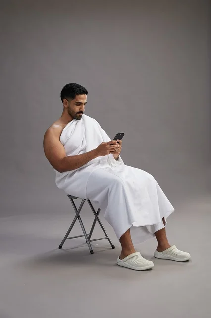 A portrait of a Saudi pilgrim in Ihram clothing using a mobile phone and sitting on a chair, preparing to perform the rituals of Hajj and Umrah, with a gray background.