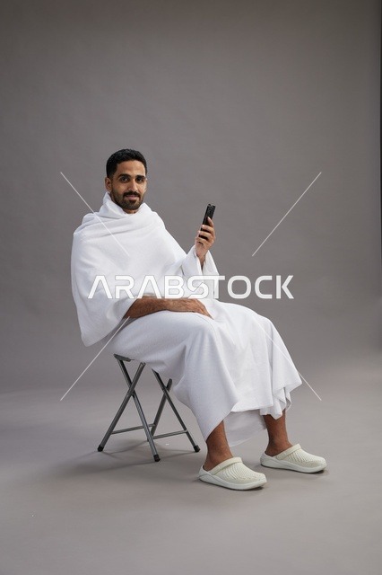 بورتريه لحاج سعودي بملابس الإحرام يستخدم الهاتف المحمول ويجلس على كرسي, الاستعداد لاداء مناسك الحج والعمره, خلفيه رماديه