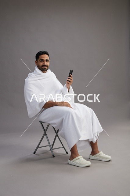 بورتريه لحاج سعودي بملابس الإحرام يستخدم الهاتف المحمول ويجلس على كرسي, الاستعداد لاداء مناسك الحج والعمره, خلفيه رماديه