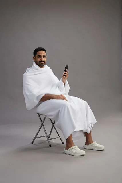 A portrait of a Saudi pilgrim in Ihram clothing using a mobile phone and sitting on a chair, preparing to perform the rituals of Hajj and Umrah, with a gray background.