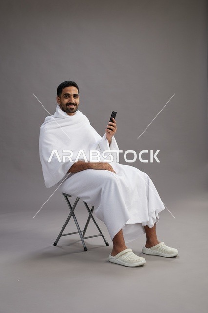 بورتريه لحاج سعودي بملابس الإحرام يستخدم الهاتف المحمول ويجلس على كرسي, الاستعداد لاداء مناسك الحج والعمره, خلفيه رماديه