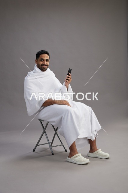 بورتريه لحاج سعودي بملابس الإحرام يستخدم الهاتف المحمول ويجلس على كرسي, الاستعداد لاداء مناسك الحج والعمره, خلفيه رماديه