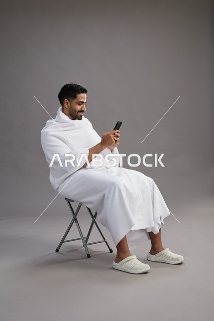 بورتريه لحاج سعودي بملابس الإحرام يستخدم الهاتف المحمول ويجلس على كرسي, الاستعداد لاداء مناسك الحج والعمره, خلفيه رماديه