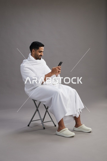 بورتريه لحاج سعودي بملابس الإحرام يستخدم الهاتف المحمول ويجلس على كرسي, الاستعداد لاداء مناسك الحج والعمره, خلفيه رماديه
