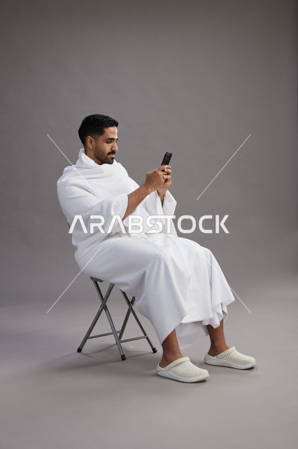 A portrait of a Saudi pilgrim in Ihram clothing using a mobile phone and sitting on a chair, preparing to perform the rituals of Hajj and Umrah, with a gray background.