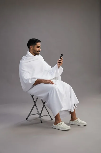 A portrait of a Saudi pilgrim in Ihram clothing using a mobile phone and sitting on a chair, preparing to perform the rituals of Hajj and Umrah, with a gray background.