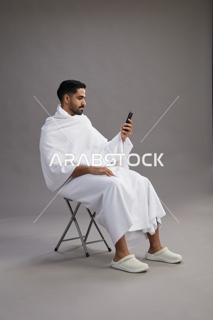 بورتريه لحاج سعودي بملابس الإحرام يستخدم الهاتف المحمول ويجلس على كرسي, الاستعداد لاداء مناسك الحج والعمره, خلفيه رماديه